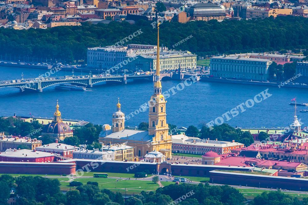 На фотографии запечатлена Петропавловская крепость, её ярким акцентом является Петропавловский собор с золотым шпилем. На заднем плане видны река Нева и Троицкий мост, соединяющий разные районы города. Исторические здания и зелёные зоны создают гармоничный пейзаж. Летний день подчёркивает красоту и величие архитектурного ансамбля Санкт-Петербурга.